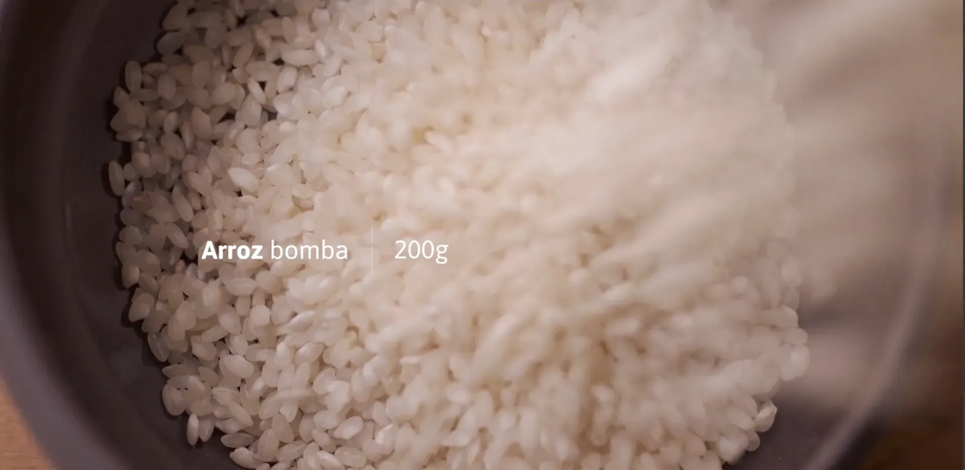 Fotograma del vídeo: primer plano de arroz cayendo en un cuenco, con el rótulo «Arroz bomba 200g»