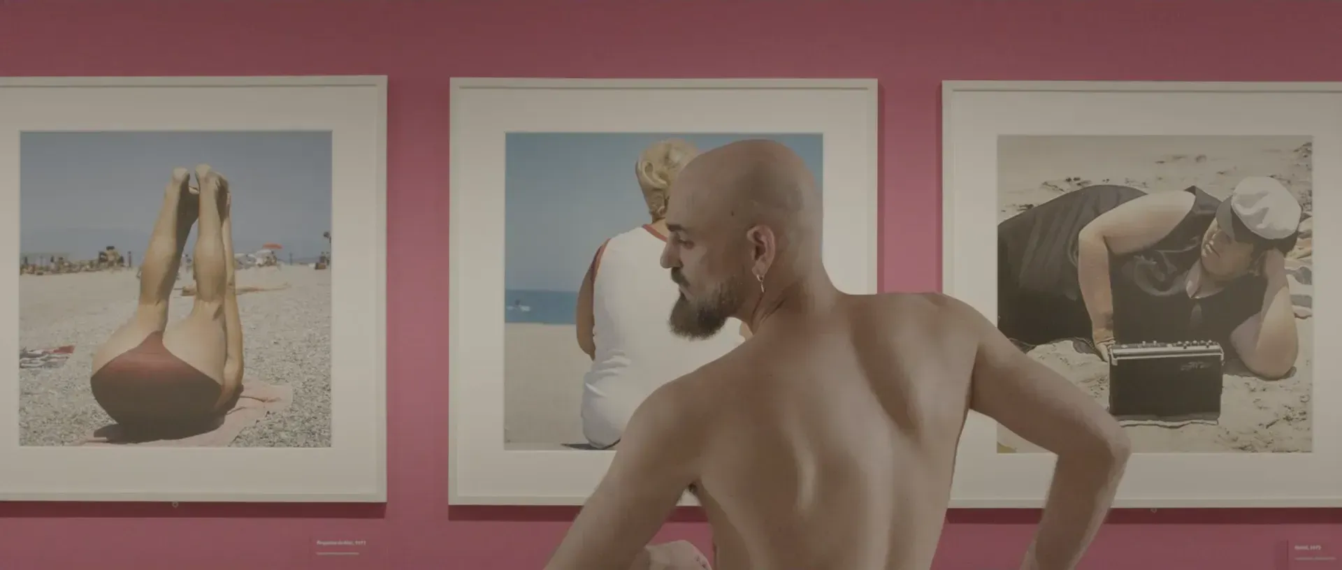 Fotograma del vídeo: un bailarín con la cabeza afeitada y barba, con el torso desnudo, visto de espaldas y de perfil, entre tres fotografías de playa colgadas en una pared rosa