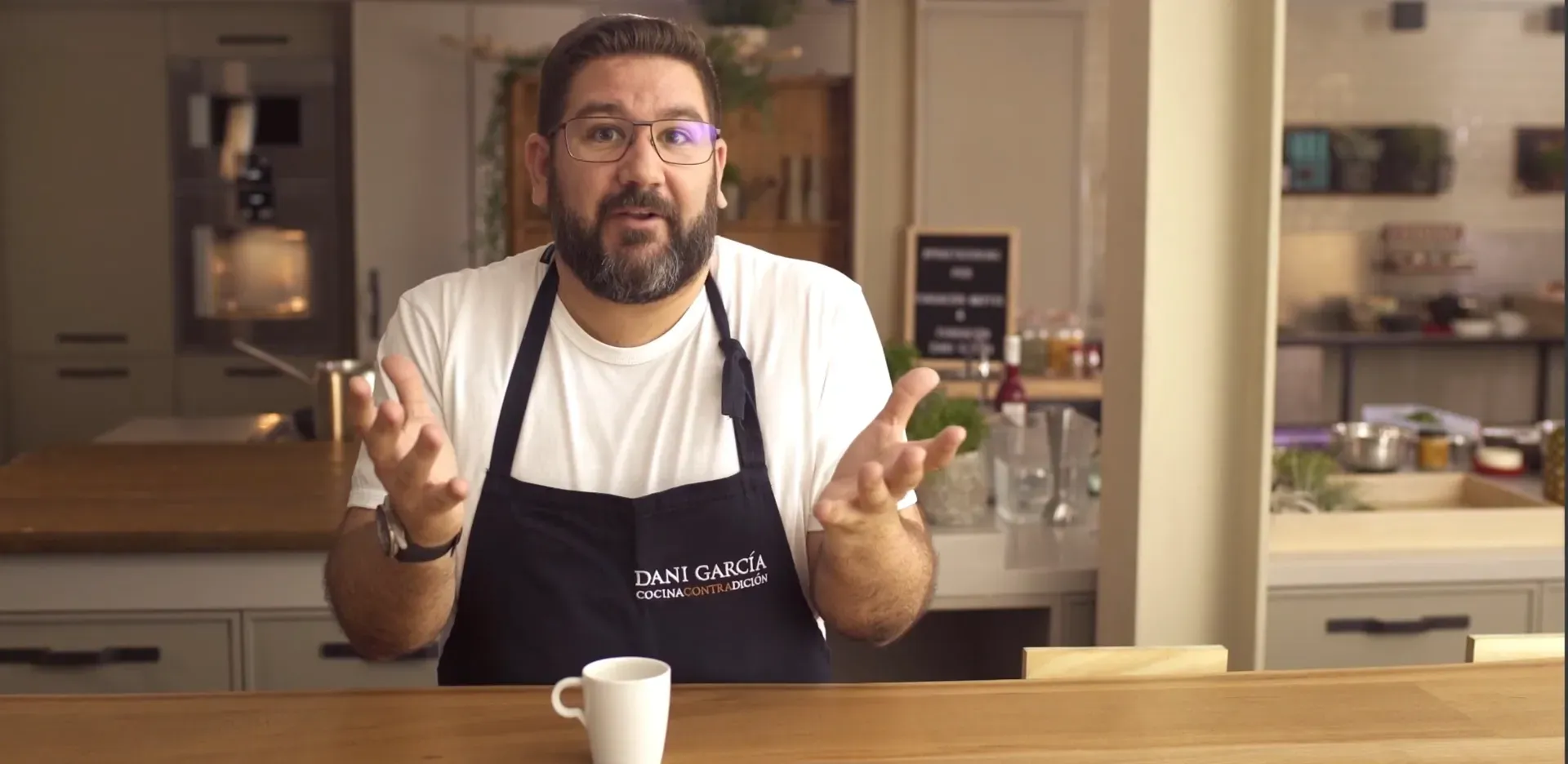 Fotograma del vídeo: Dani García, con delantal azul marino, habla a cámara con las manos abiertas desde la mesa de una cocina, con una taza delante
