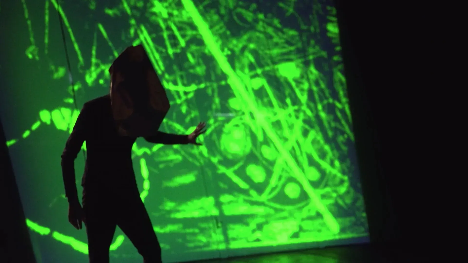 Fotograma del vídeo: la silueta de una persona con una bolsa de papel en la cabeza ante una proyección de trazos verdes fluorescentes, con una mano levantada hacia la imagen