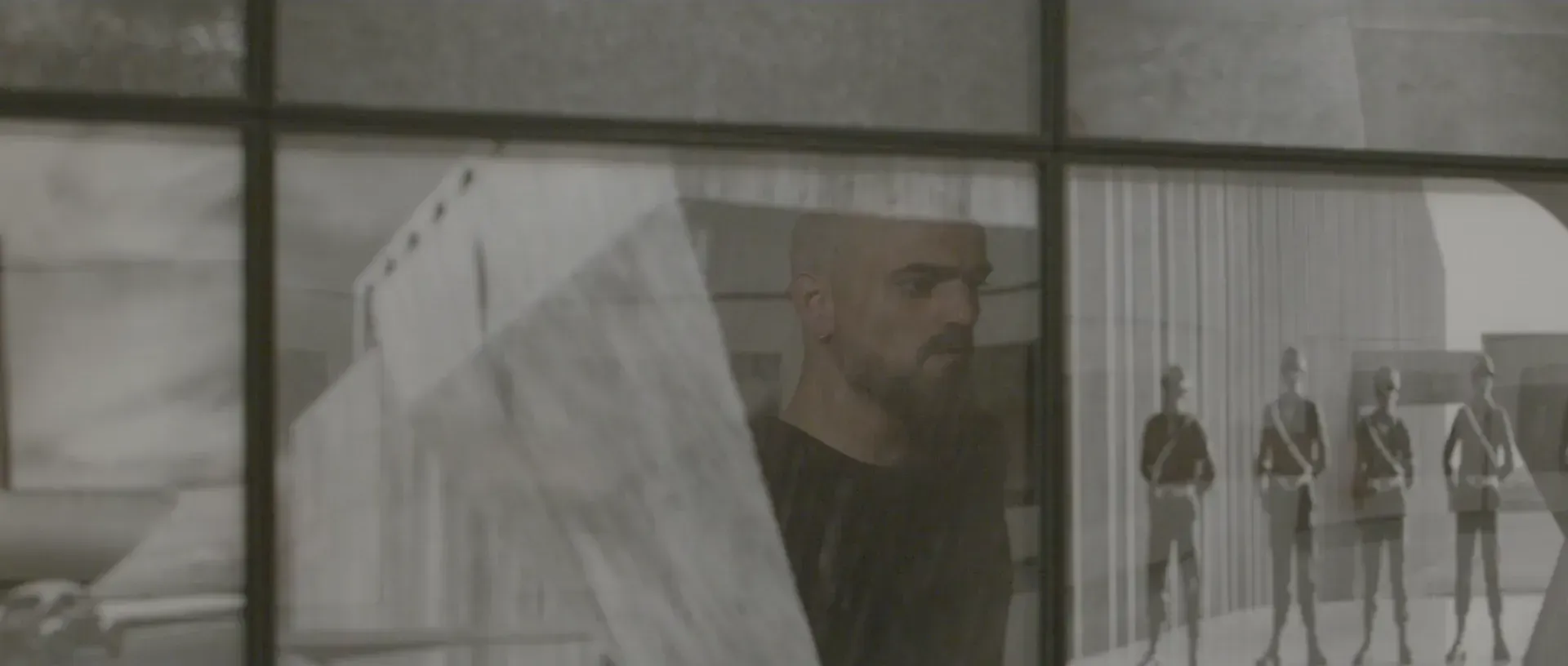 Fotograma del vídeo: en blanco y negro, el rostro del bailarín, con barba y un pendiente, superpuesto a una fotografía de figuras uniformadas en fila, tras unas líneas de marco