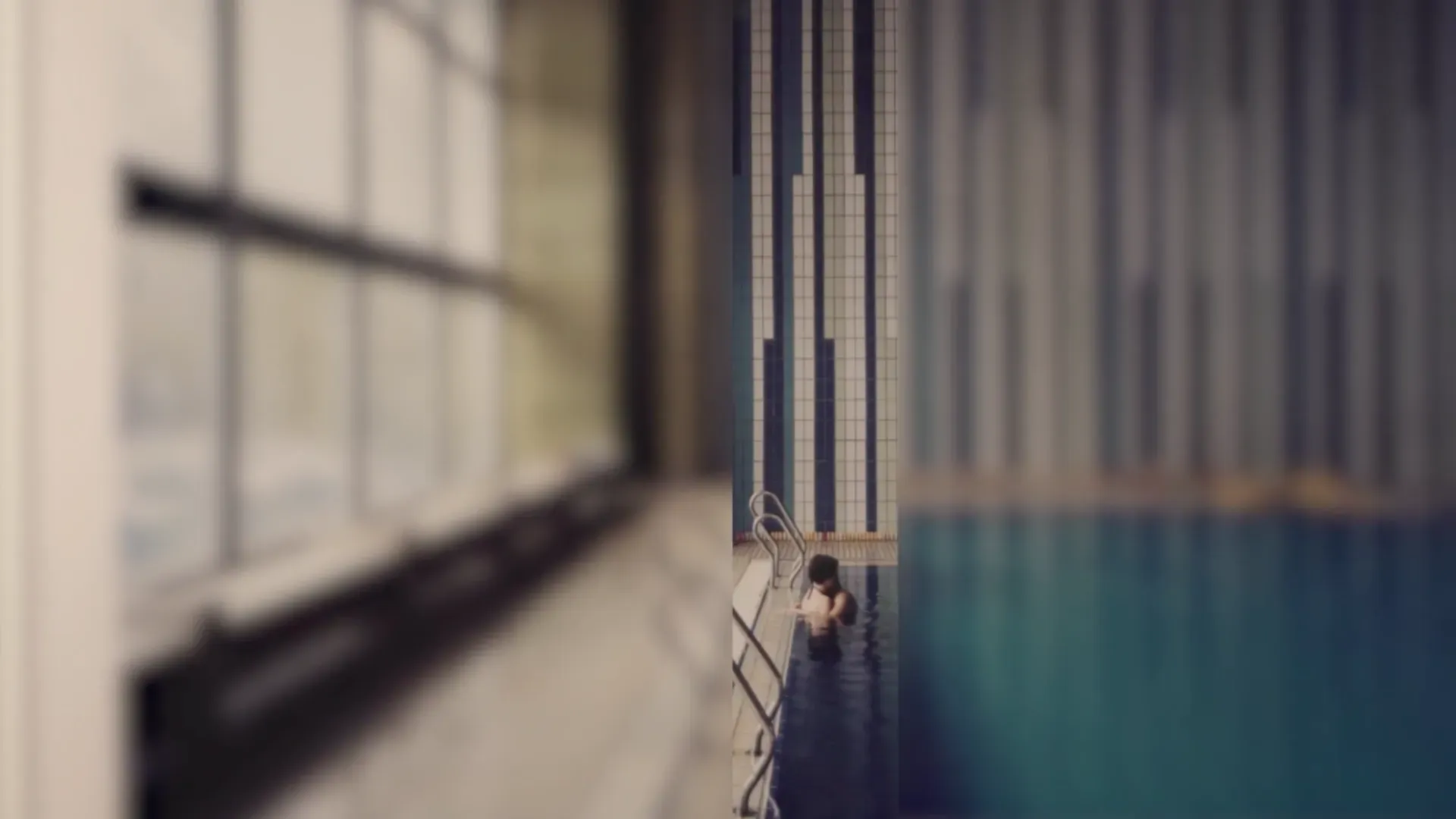 Fotograma del vídeo: una piscina cubierta con azulejos blancos y azules; una persona asoma en el agua junto a una escalera, en una tira vertical nítida entre dos zonas desenfocadas