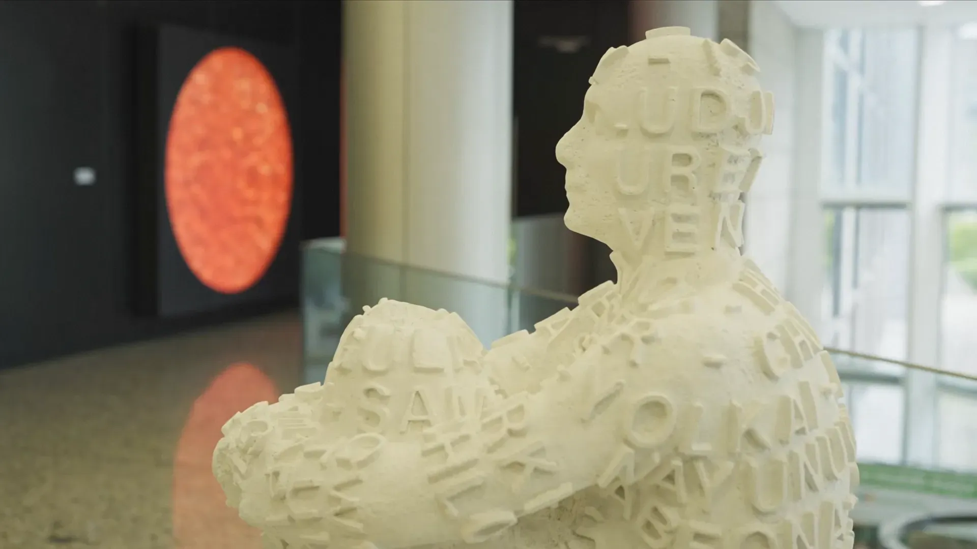 Fotograma del vídeo: una escultura blanca de una figura sentada hecha de letras, en un vestíbulo acristalado, con una obra circular roja al fondo